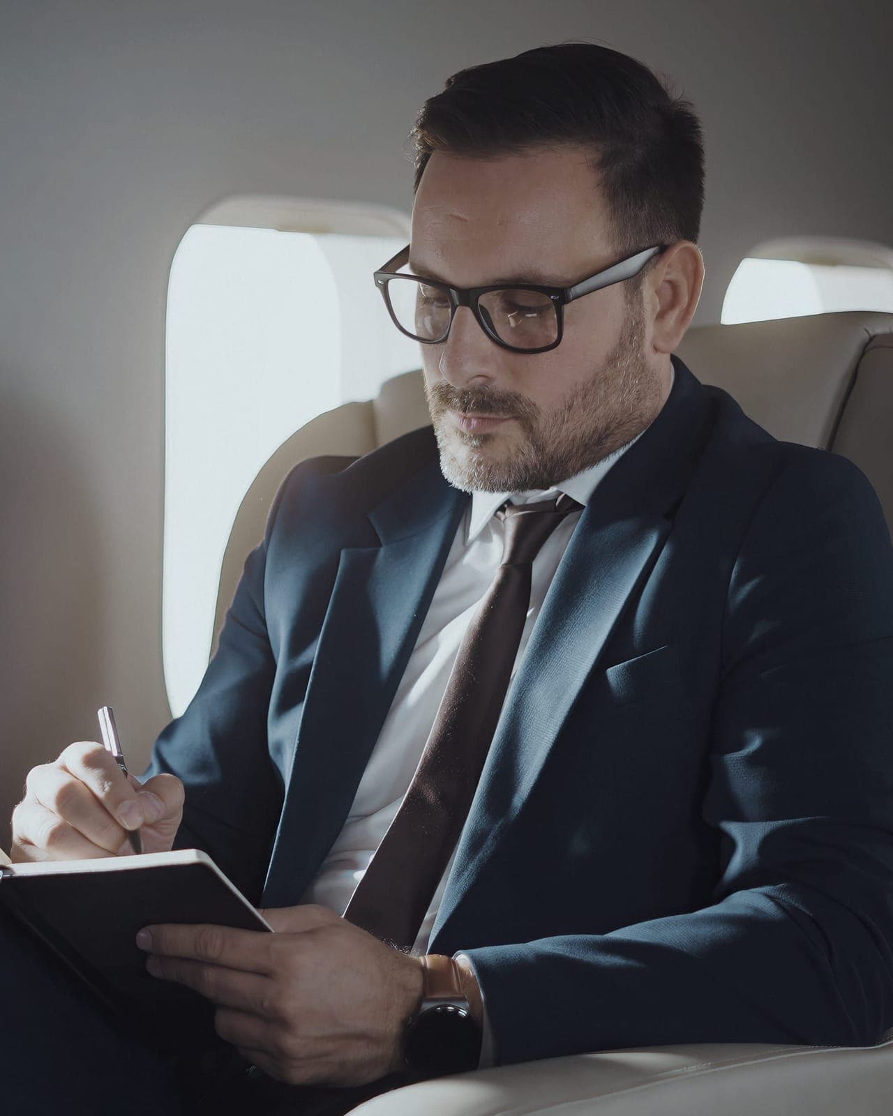 Geschäftsmann im dunkelblauen Anzug mit Brille, an Bord eines Privatjets, macht Notizen in ein Notizbuch.